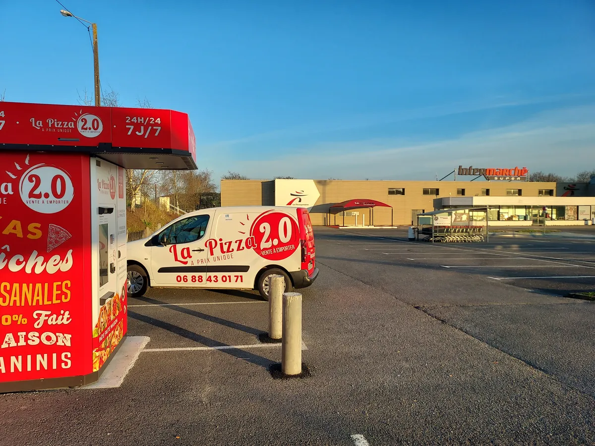 Le distributeur de pizzas La Pizza 2.0 de Châteaulin, en bordure du parking d'Intermarché, avec la camionnette de la maison garée à côté et le magasin au fond